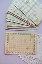 Close-up of the front of the postcard featuring the yellow and white wide gingham background with the line-art sketches of a basket, duck, and flowers, and the quote, "A lot of little things make my heart happy."