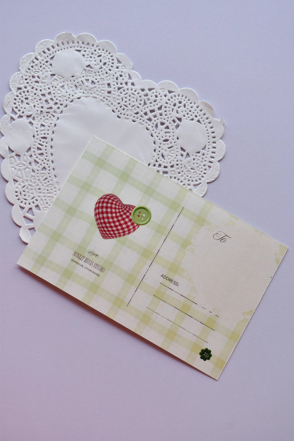 A horizontal postcard reverse side with a pale green and white wide plaid pattern. It features a red gingham patchwork heart secured with a green button, resting on a white heart-shaped lace doily.