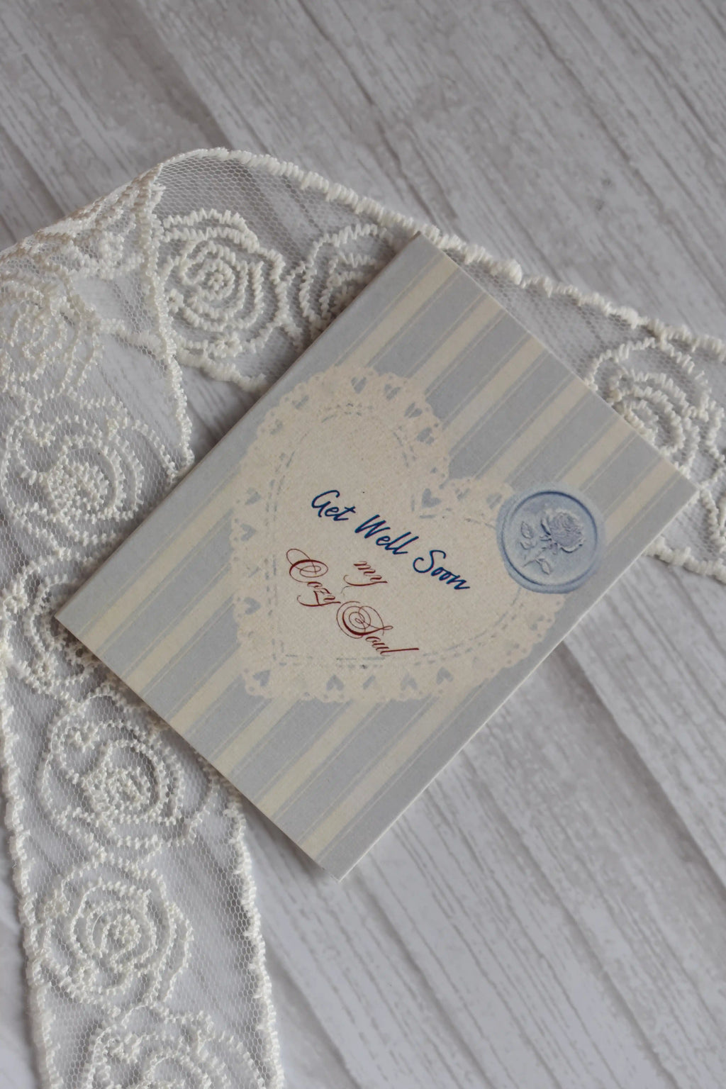 Angled flat lay of the Sunlit Hugs Get Well Soon Cozy Soul Greeting Card showing the light blue stripes, lace heart, and faux wax seal on a wooden background next to white lace.