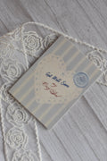 Angled flat lay of the Sunlit Hugs Get Well Soon Cozy Soul Greeting Card showing the light blue stripes, lace heart, and faux wax seal on a wooden background next to white lace.