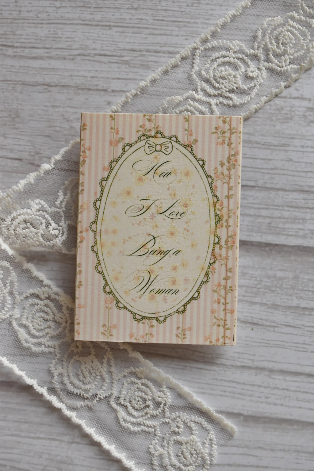 Angled flat lay of the Sunlit Hugs How I Love Being a Woman Greeting Card showing the vintage pink striped and floral design resting beside decorative white lace.