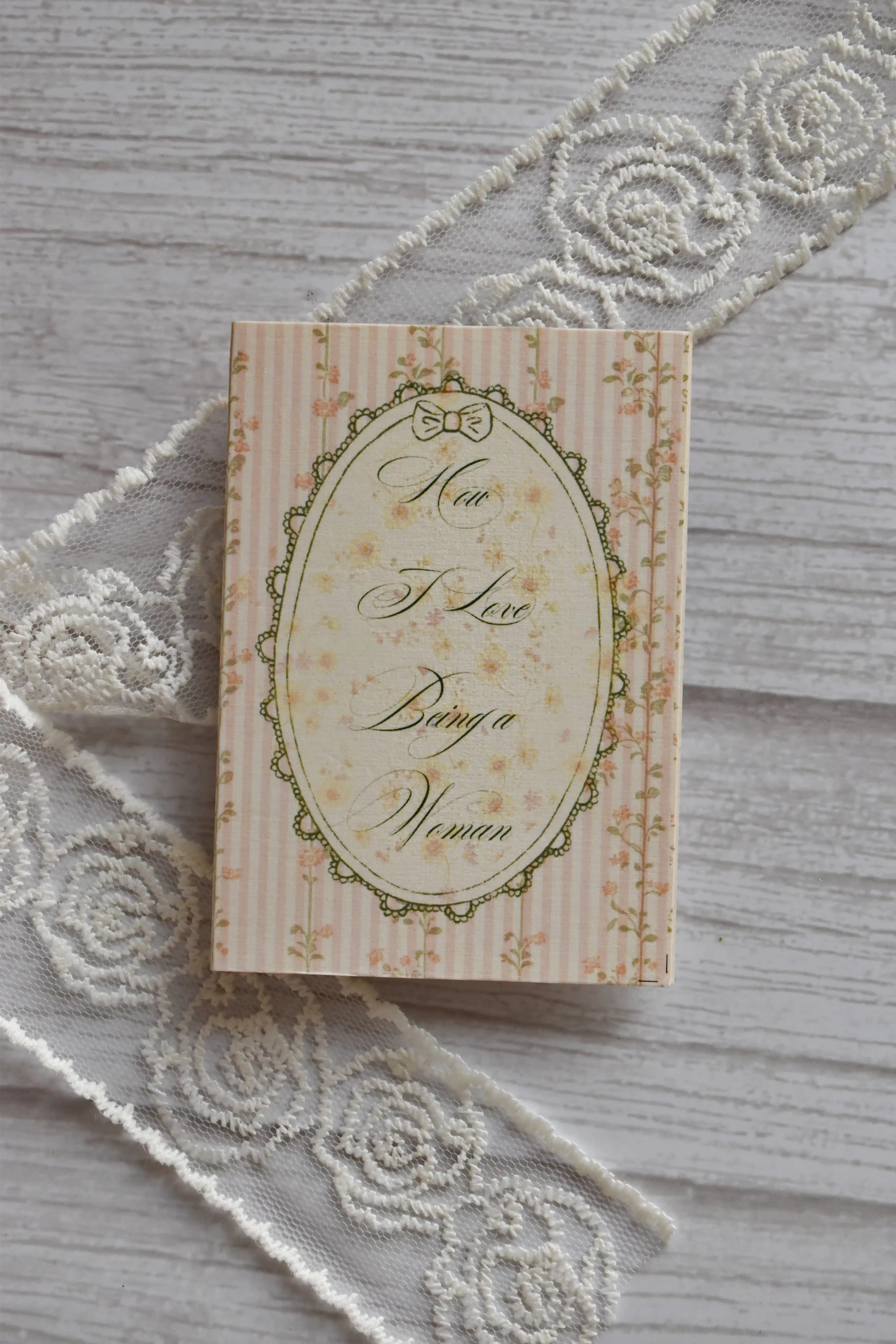 Angled flat lay of the Sunlit Hugs How I Love Being a Woman Greeting Card showing the vintage pink striped and floral design resting beside decorative white lace.