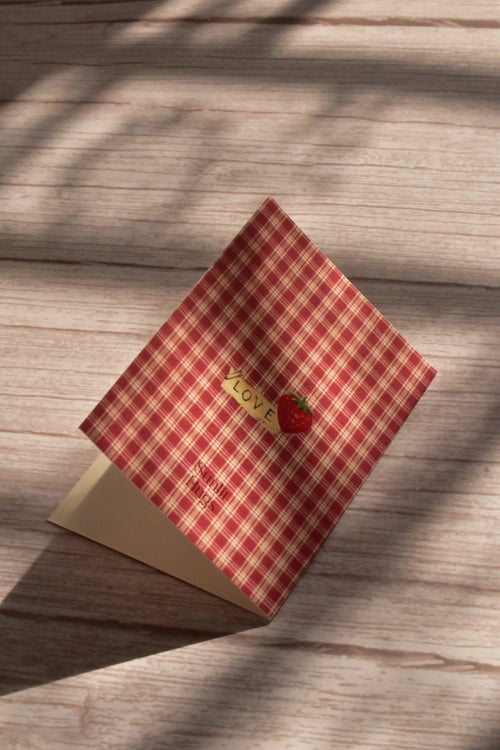 Standing angled view of the Sunlit Hugs Mi Amor Por Ti Greeting Card bathed in warm sunlight, showcasing the rustic red plaid design and vintage romantic details.