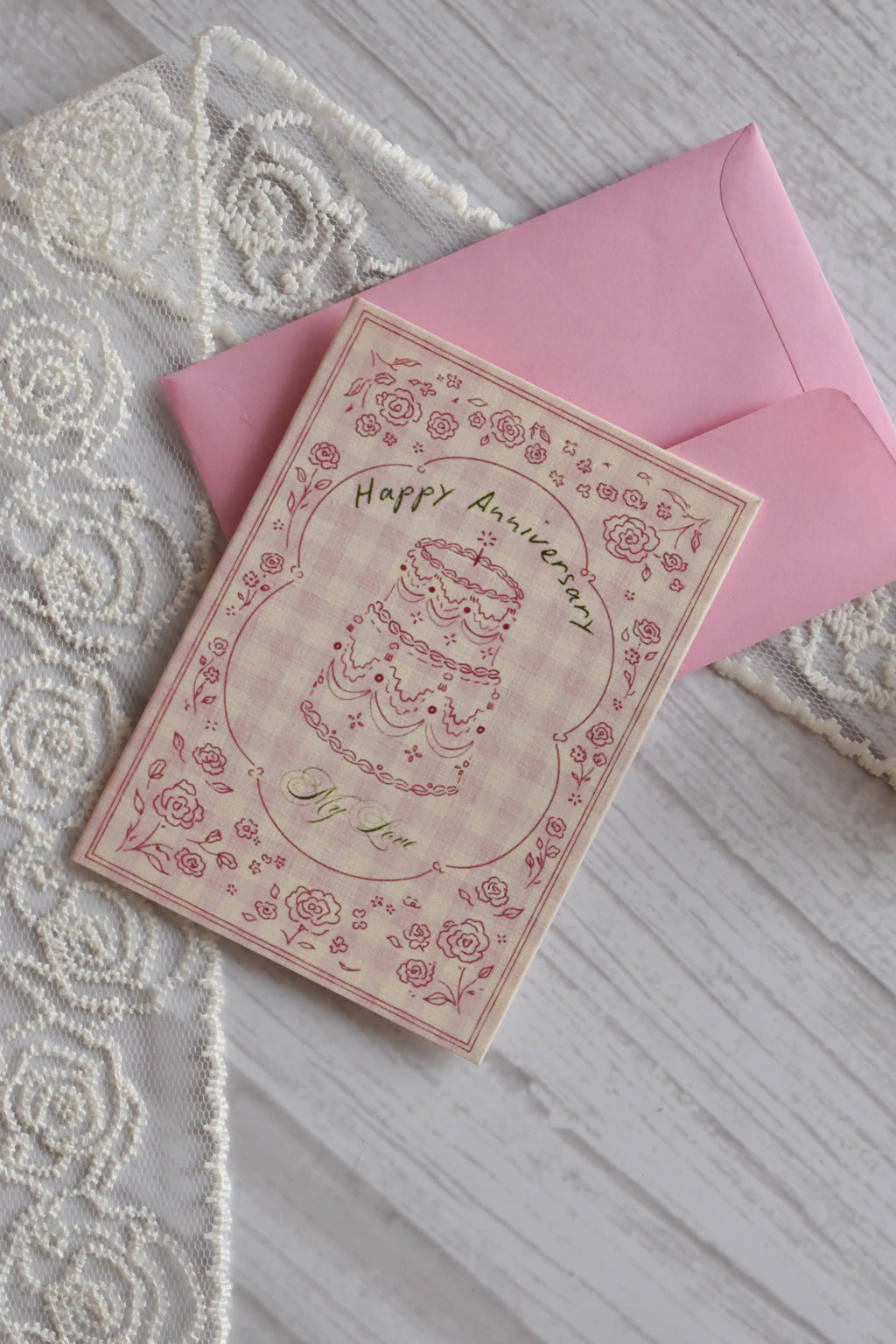 Angled flat lay of the Sunlit Hugs Happy Anniversary Vintage Cake Greeting Card placed next to decorative white floral lace.