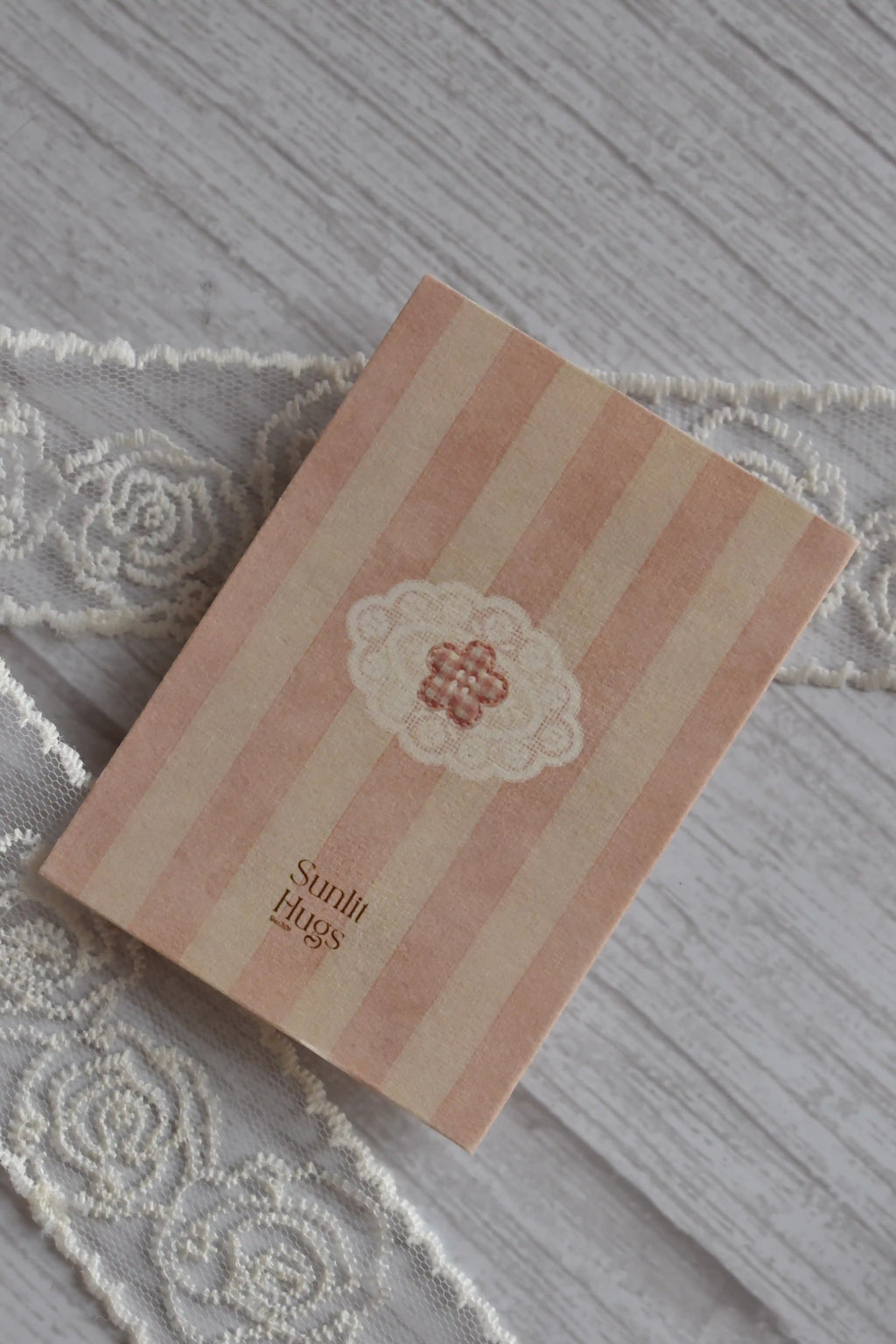 Back cover of the Sunlit Hugs Life Feels Kinder With You Greeting Card featuring pink stripes, a small lace and floral emblem, and the brand logo.
