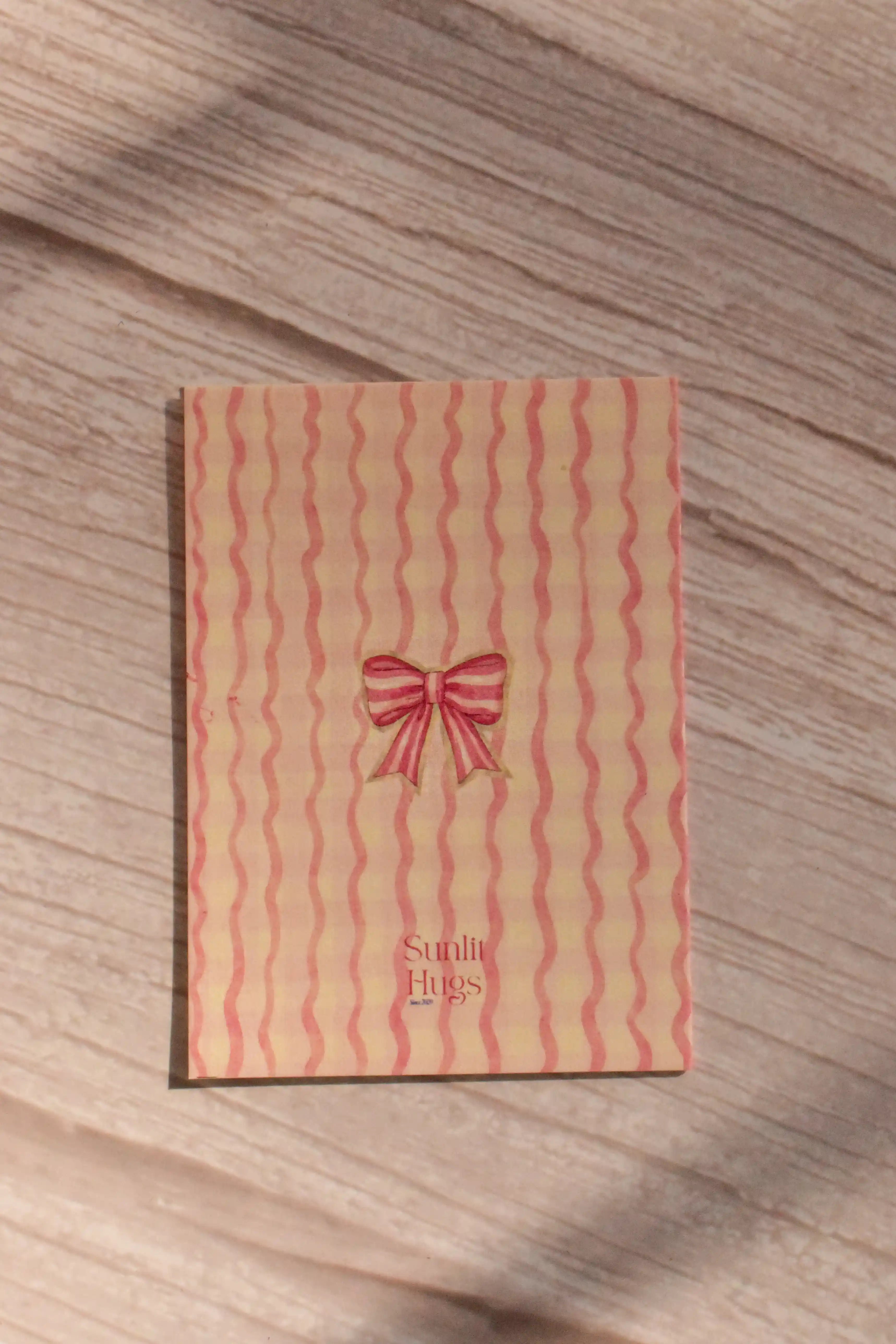 Back cover of the Sunlit Hugs Your Heart Is My Home Greeting Card showing a wavy pink striped pattern, a delicate watercolor red bow, and the Sunlit Hugs logo.