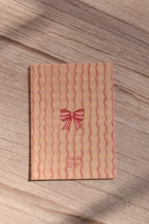 Back cover of the Sunlit Hugs Your Heart Is My Home Greeting Card showing a wavy pink striped pattern, a delicate watercolor red bow, and the Sunlit Hugs logo.
