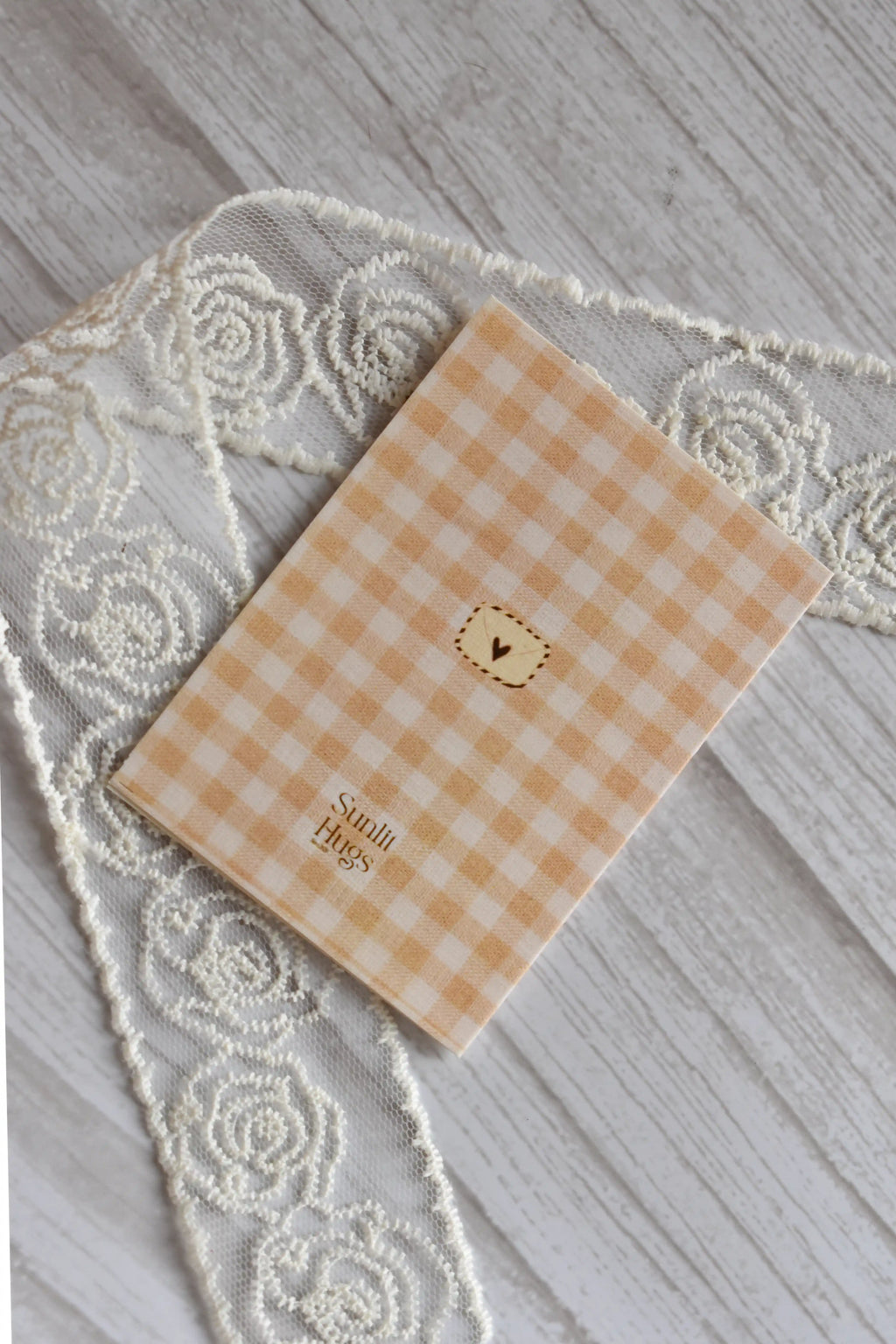 Back of the Sunlit Hugs Happy Birthday Sock Twin Greeting Card featuring a beige gingham pattern, a small heart envelope illustration, and the brand logo, placed next to white lace.