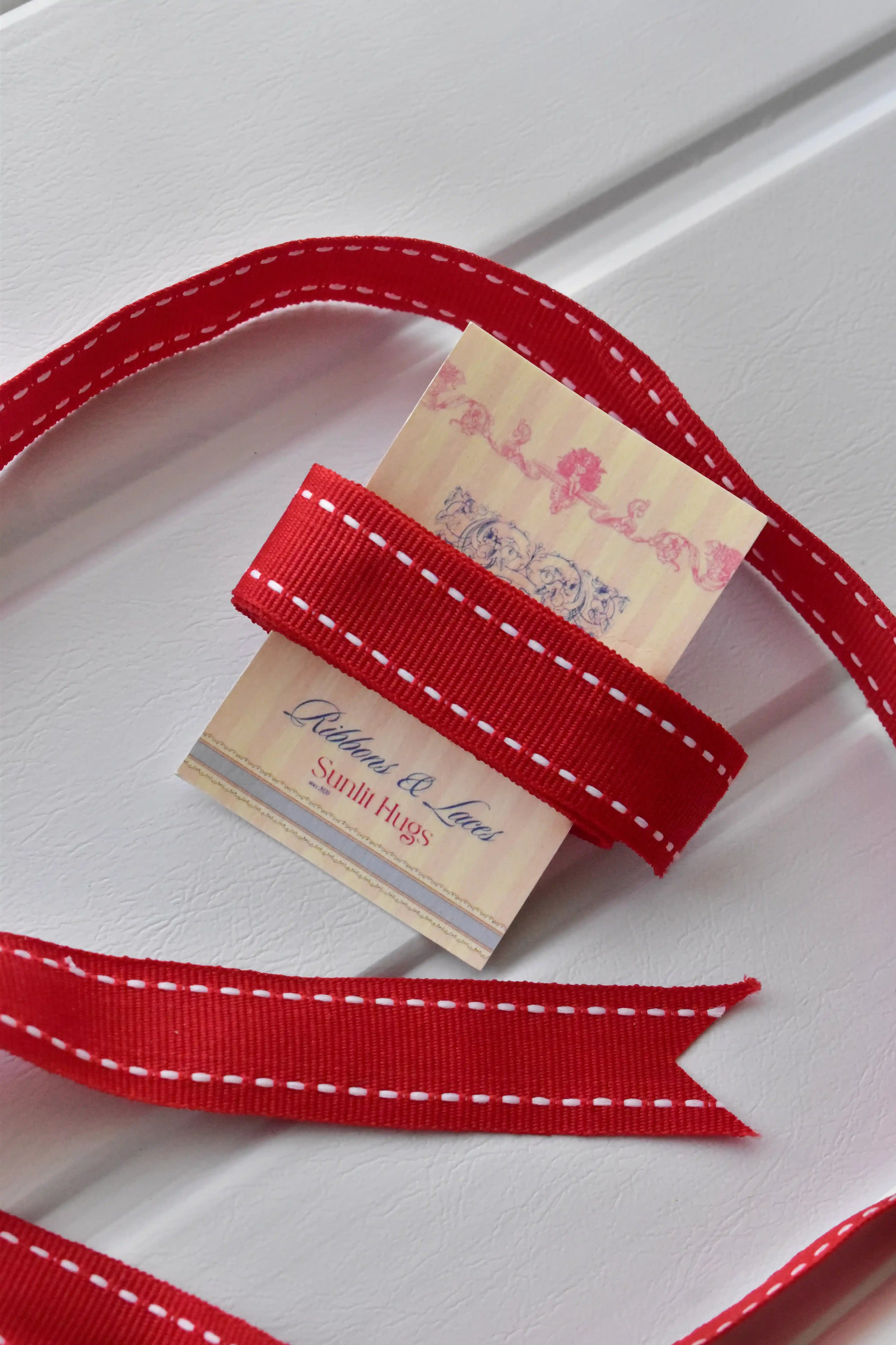 A detail shot showing a loose section of the Berry Stitch ribbon wrapped around a "Sunlit Hugs" tag. The image focuses on the ribbon's bright red color and prominent white dashed stitching.