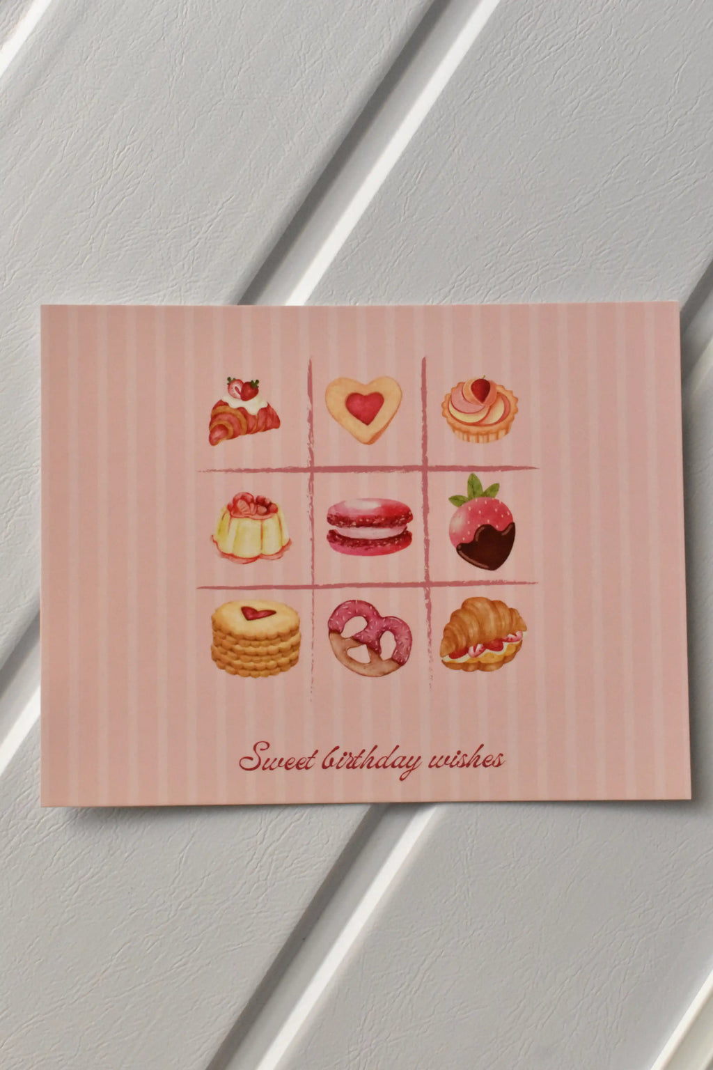 This image showcases the main front design of the Birthday Bakes Postcard, highlighting the vibrant grid of nine whimsical desserts and the soft pink striped background.