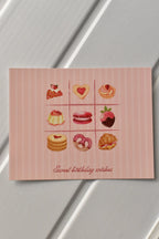This image showcases the main front design of the Birthday Bakes Postcard, highlighting the vibrant grid of nine whimsical desserts and the soft pink striped background.