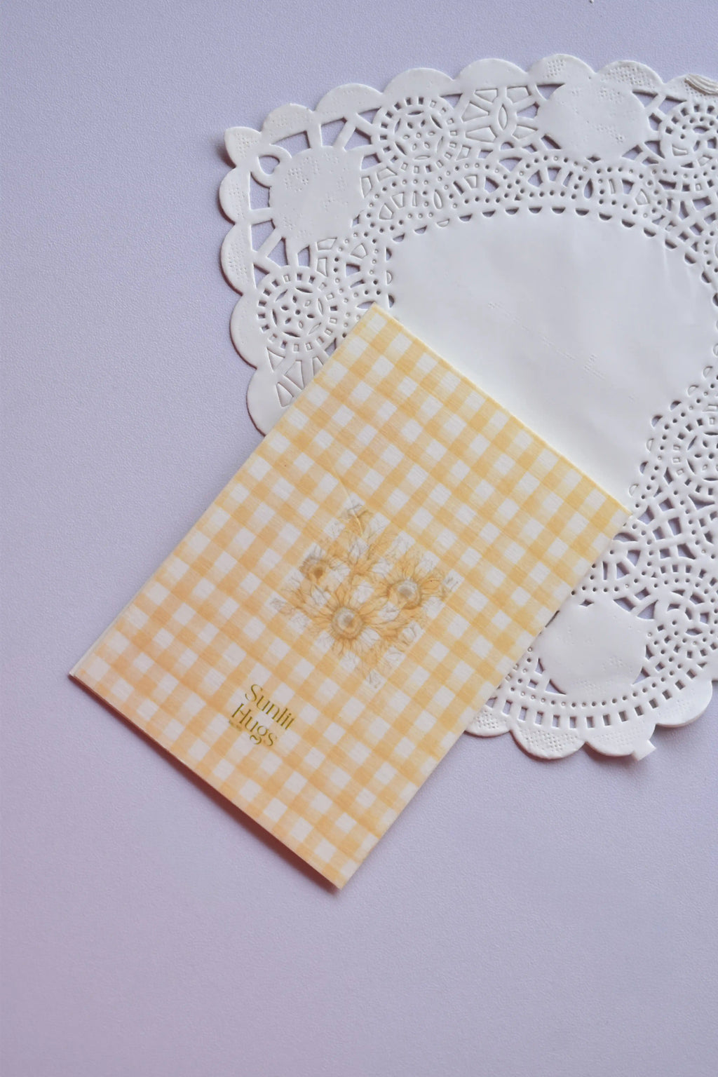 Detailed view of a vertical greeting card with a yellow and white gingham pattern. It features a tall sunflower stem and a decorative strip of printed lace down the left side. The text reads "Wishing you Birthday Sunshine." It is set against a white paper doily.