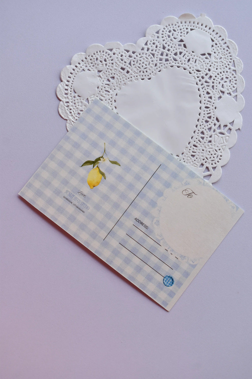 The back view of a postcard resting on a white heart-shaped paper doily. The design is a soft blue and white wide gingham pattern, with a small lemon illustration. It includes labeled fields for "To" and "Address," and a light blue circle detail.