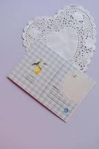 The back view of a postcard resting on a white heart-shaped paper doily. The design is a soft blue and white wide gingham pattern, with a small lemon illustration. It includes labeled fields for "To" and "Address," and a light blue circle detail.