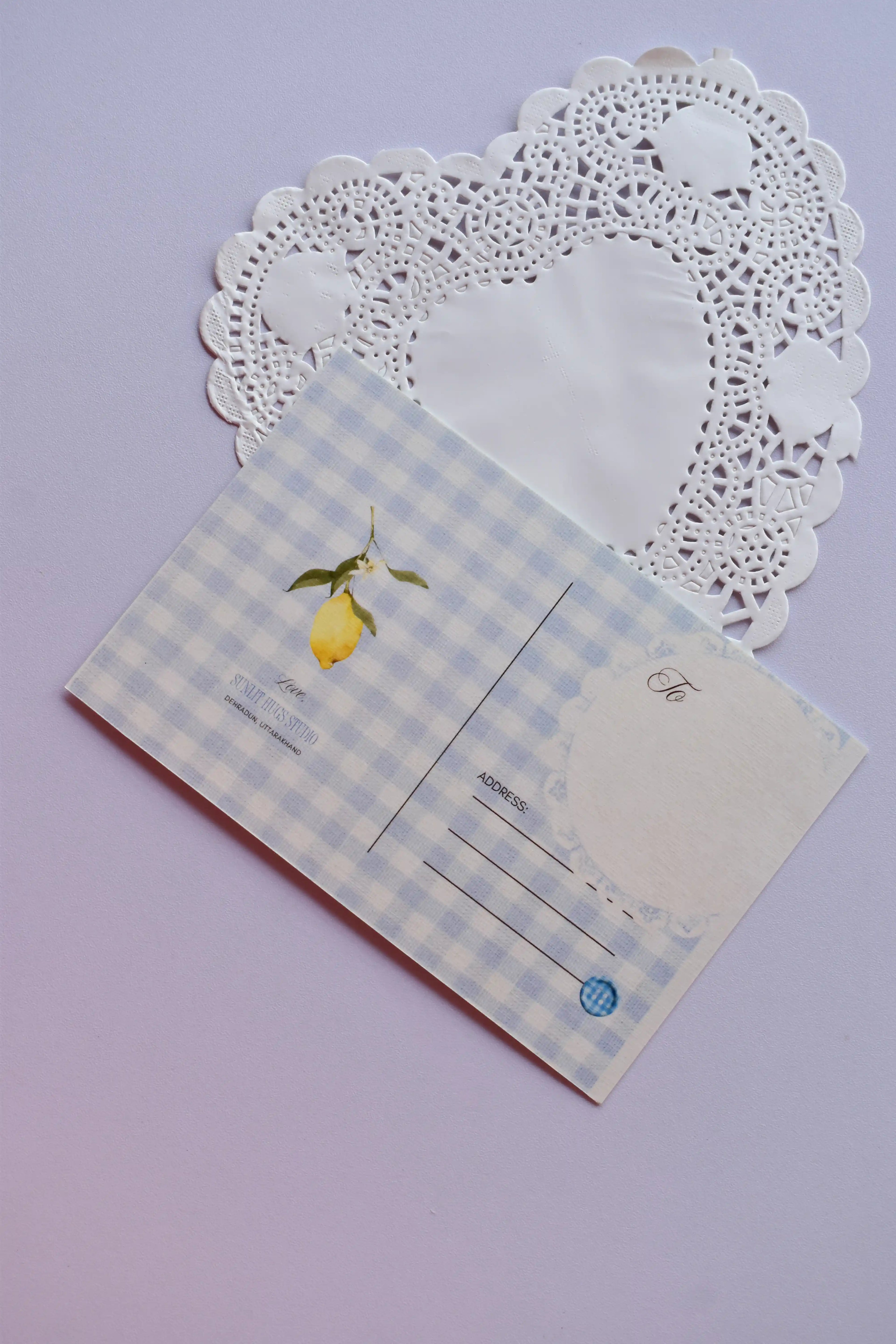 The back view of a postcard resting on a white heart-shaped paper doily. The design is a soft blue and white wide gingham pattern, with a small lemon illustration. It includes labeled fields for "To" and "Address," and a light blue circle detail.