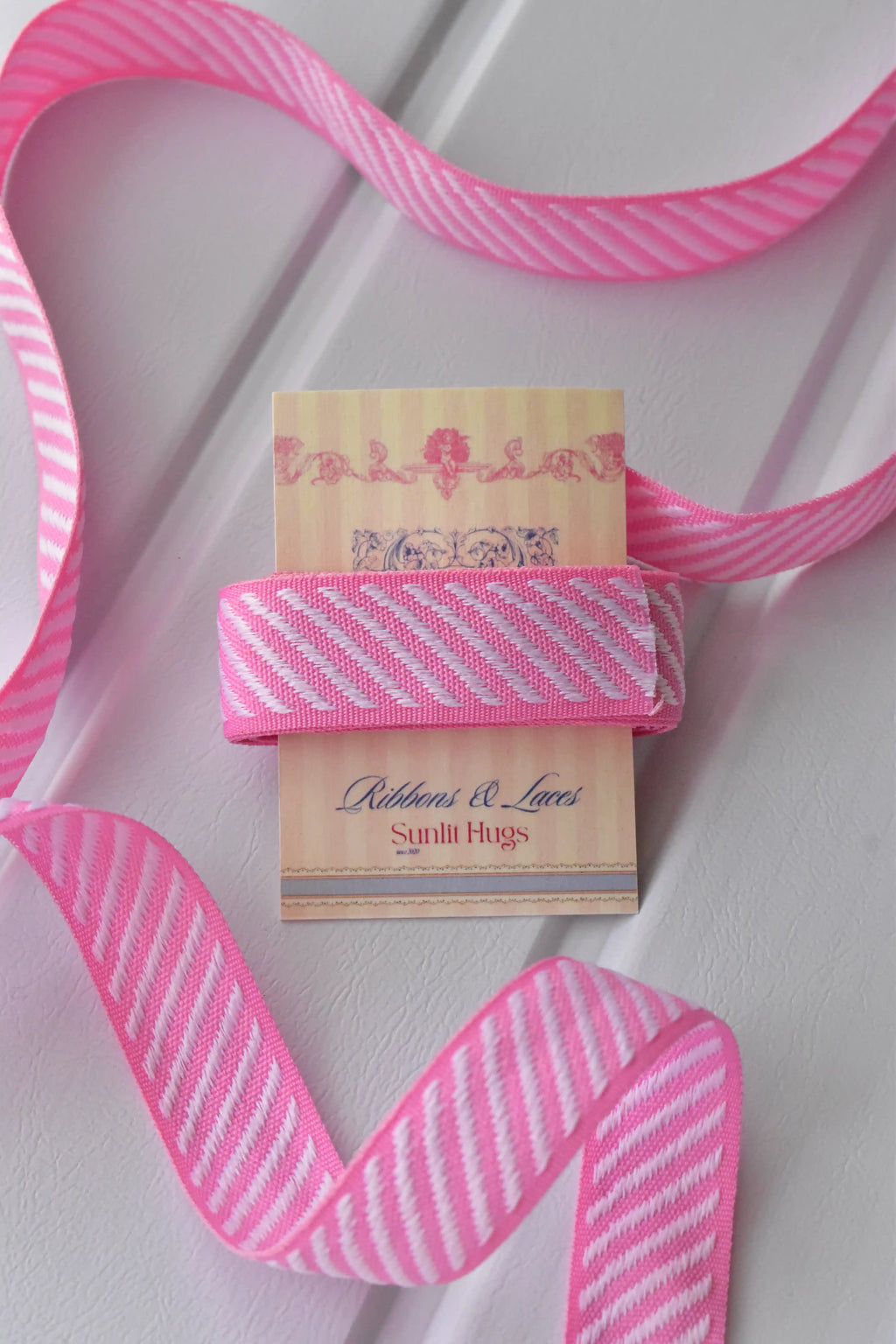 A swatch of pink grosgrain ribbon with white diagonal stripes wrapped around a Sunlit Hugs tag, with a long strand unwound.