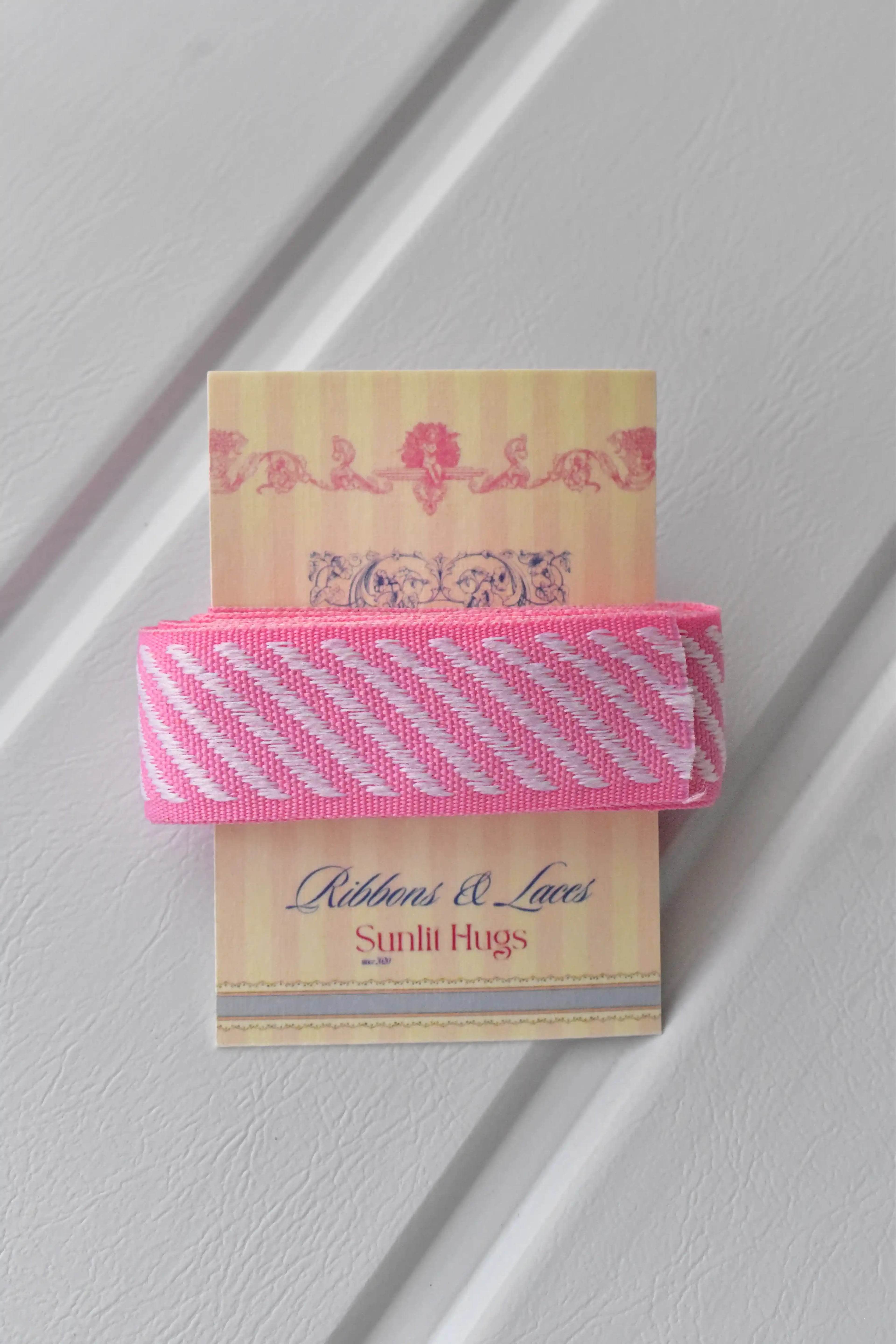 A close-up detail shot of the Candy Stripes ribbon. The ribbon is wrapped around the "Sunlit Hugs Ribbons & Laces" brand tag, showcasing its texture and the crispness of its pink and white diagonal stripes.