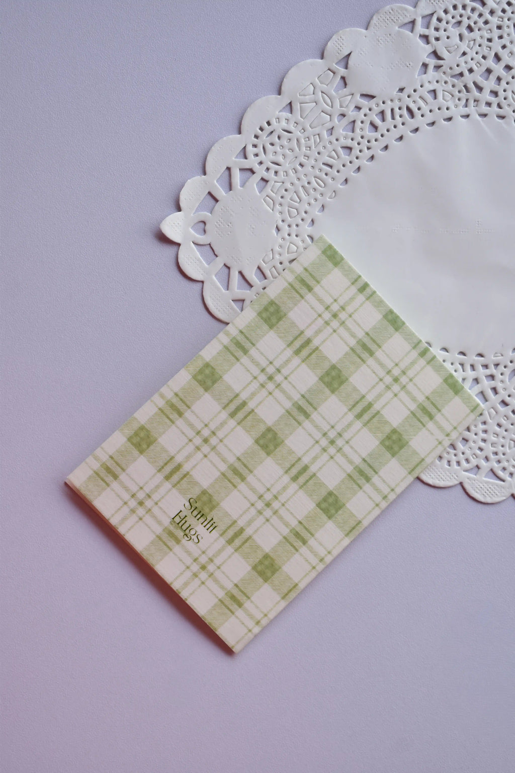 Close-up of a small greeting card featuring a soft pale green and white gingham plaid pattern. The text "Sunlit Hugs" is printed on the bottom left corner. The card rests on a white lace paper doily.