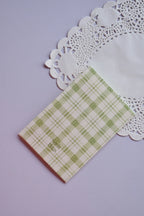 Close-up of a small greeting card featuring a soft pale green and white gingham plaid pattern. The text "Sunlit Hugs" is printed on the bottom left corner. The card rests on a white lace paper doily.