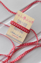 This image features the Cherry Picnic narrow ribbon with a long, flowing strand unravelled, showcasing its full red and white checkered pattern and texture. A small section is also wrapped around a "Sunlit Hugs" brand tag.