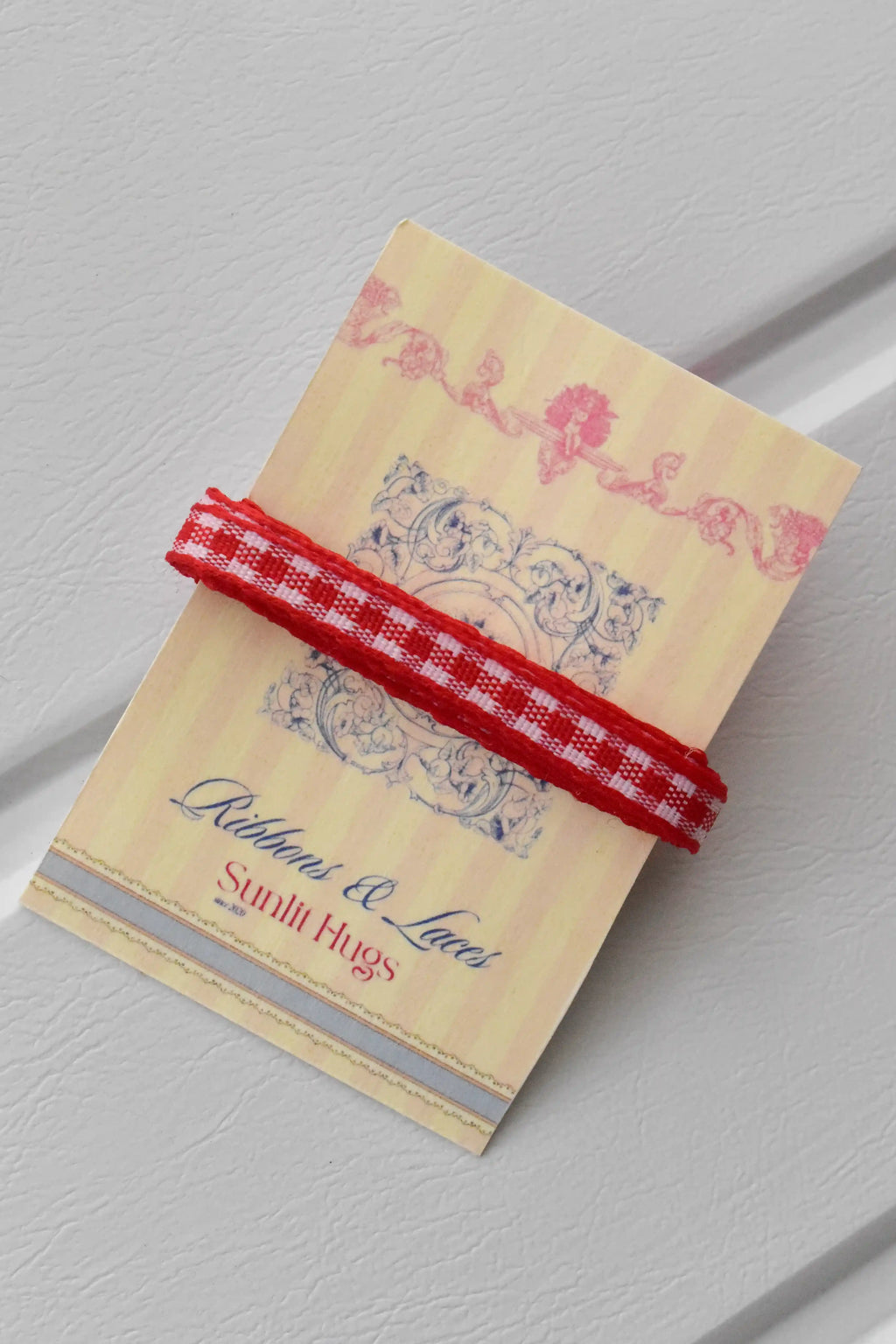 This close-up shows a swatch of the Cherry Picnic narrow ribbon, highlighting its classic red and white checkered pattern and woven texture, neatly wrapped around a "Sunlit Hugs" brand tag.
