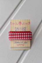 A detailed, head-on view of the Cherry Picnic Red Wide ribbon swatch. The ribbon is neatly wrapped around the "Sunlit Hugs" brand tag, showcasing the exact red and white checkered pattern and the ribbon's generous width.