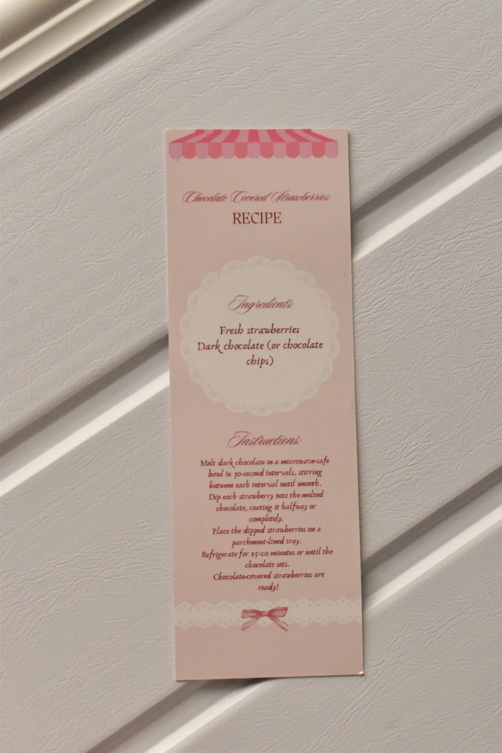 This image focuses on the practical element, displaying the detailed Chocolate Covered Strawberries Recipe printed on the back of the pastel pink bookmark, ready for kitchen inspiration.