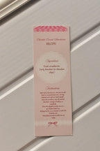 This image focuses on the practical element, displaying the detailed Chocolate Covered Strawberries Recipe printed on the back of the pastel pink bookmark, ready for kitchen inspiration.
