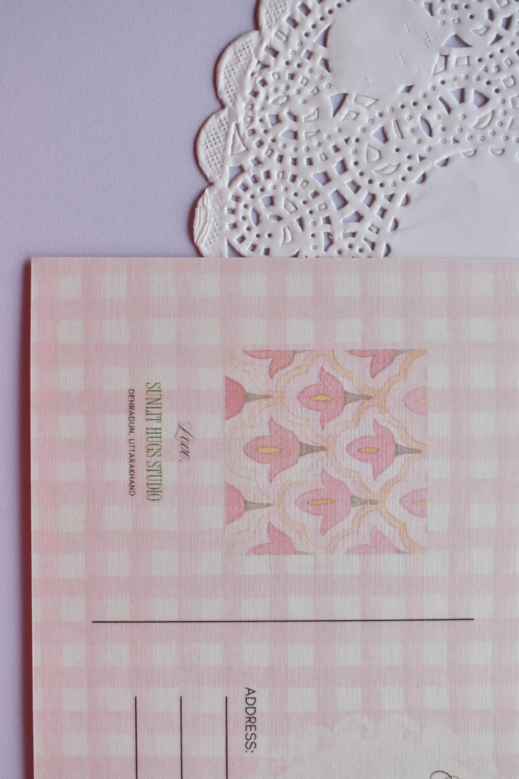 Extreme close-up of the postcard reverse, showing the pink and white wide gingham pattern and the central pink floral bell pattern square. The text "SUNLIT HUGS STUDIO" is visible.