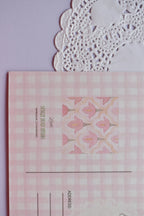 Extreme close-up of the postcard reverse, showing the pink and white wide gingham pattern and the central pink floral bell pattern square. The text "SUNLIT HUGS STUDIO" is visible.
