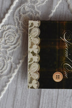 Macro close-up of the Sunlit Hugs Vintage Plaid Congratulations Greeting Card focusing on the printed white lace edge and vintage button detail against the dark plaid background.
