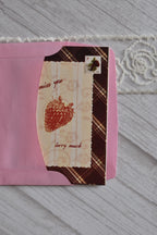 Close-up of the Sunlit Hugs I Miss You Berry Much Greeting Card inside a pink envelope, focusing on the strawberry illustration, text, and faux vintage stamp.
