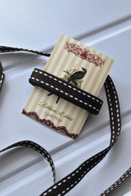 This image displays the Cocoa Stitch ribbon in full, with a long strand flowing loosely, highlighting its dark brown colour and continuous white dashed stitching. A section is also wrapped around a "Sunlit Hugs" brand tag.
