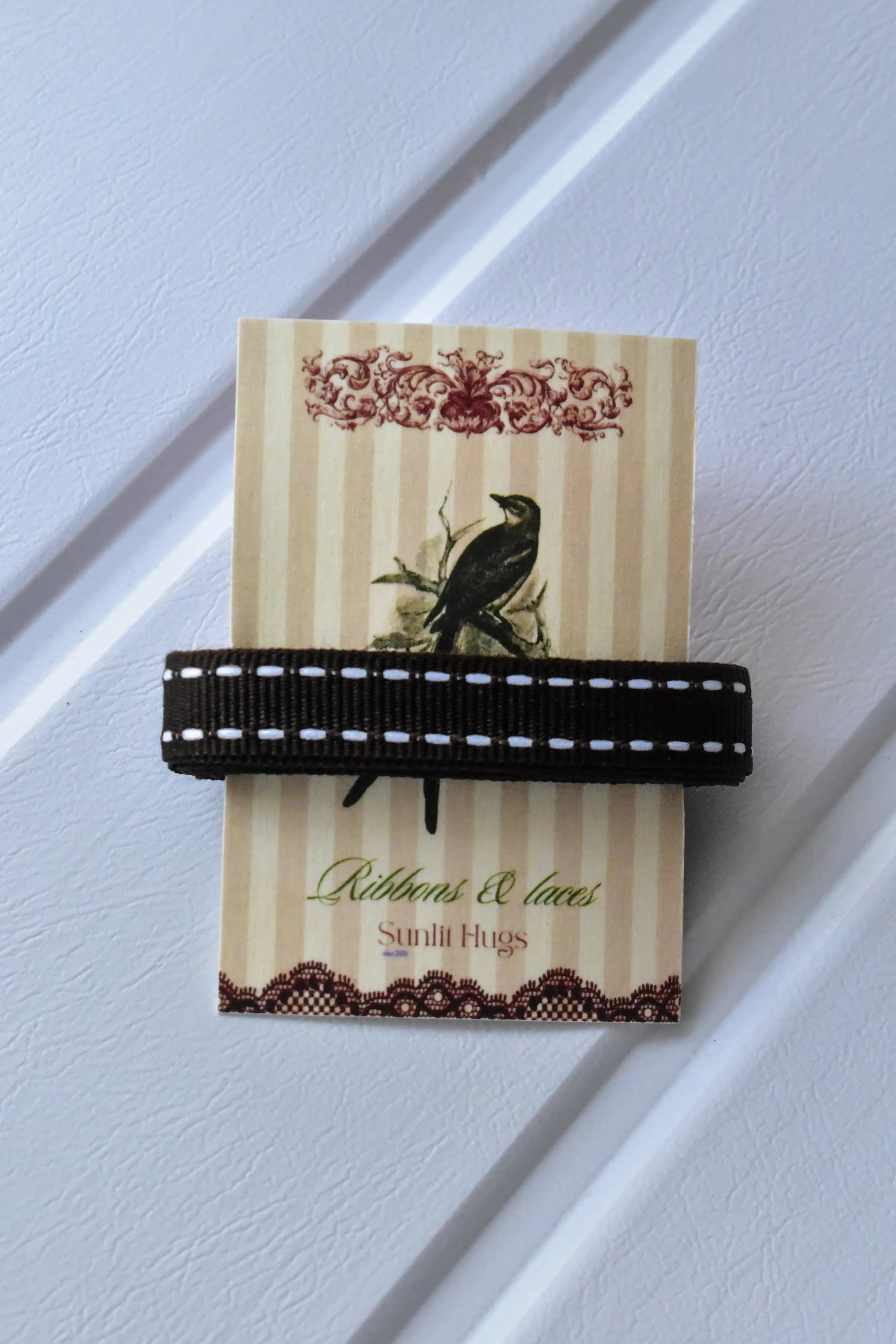 This image provides a detailed view of the Cocoa Stitch ribbon, showcasing its rich dark brown colour and the delicate white dashed stitching, neatly presented on a "Sunlit Hugs" brand tag.