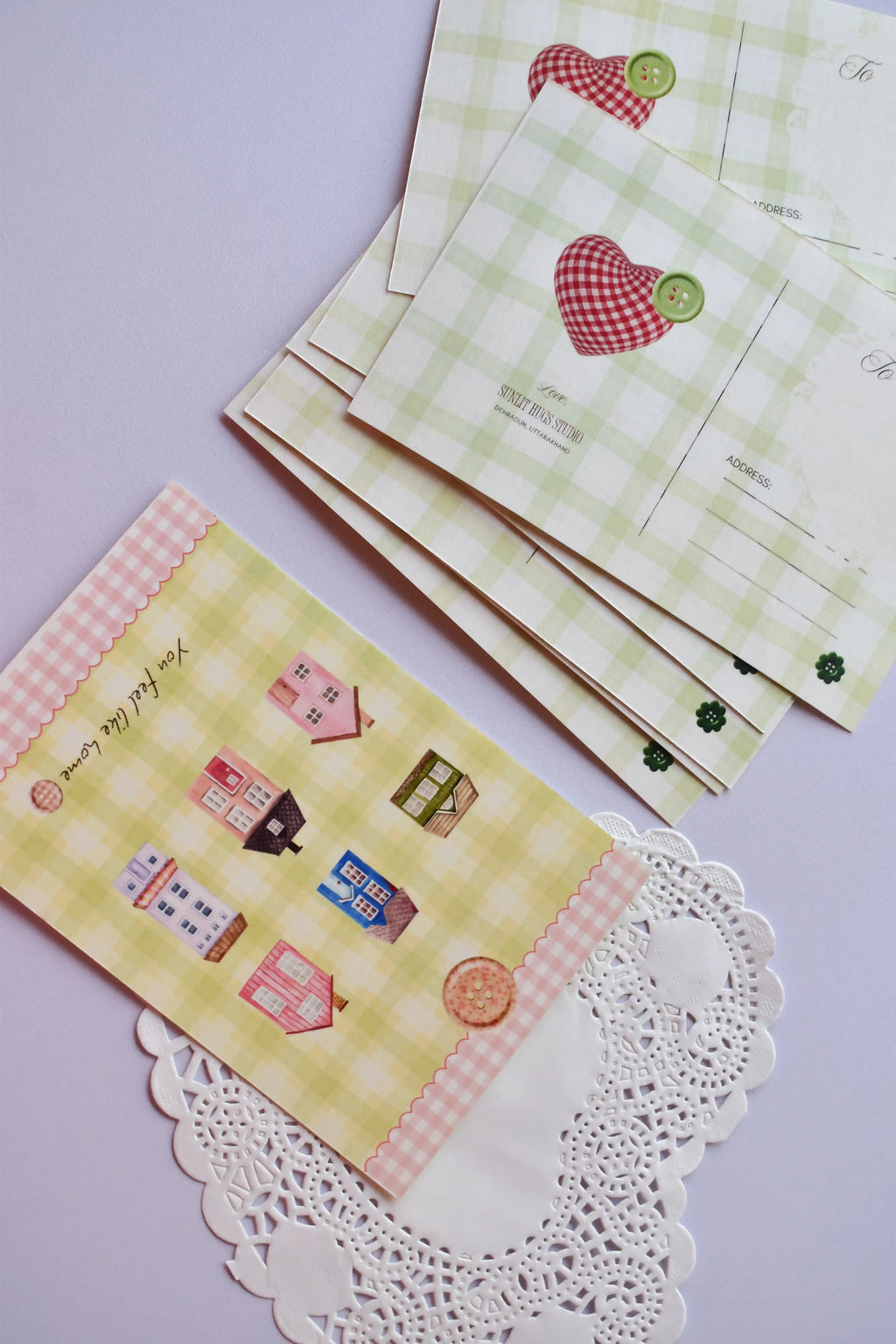 A collection of postcards showing the cozy cottage design on the bottom card, and the reverse side (or alternate card) featuring the green gingham and red patchwork heart design stacked behind it. The heart design includes spaces for address and postage.