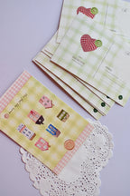 A collection of postcards showing the cozy cottage design on the bottom card, and the reverse side (or alternate card) featuring the green gingham and red patchwork heart design stacked behind it. The heart design includes spaces for address and postage.