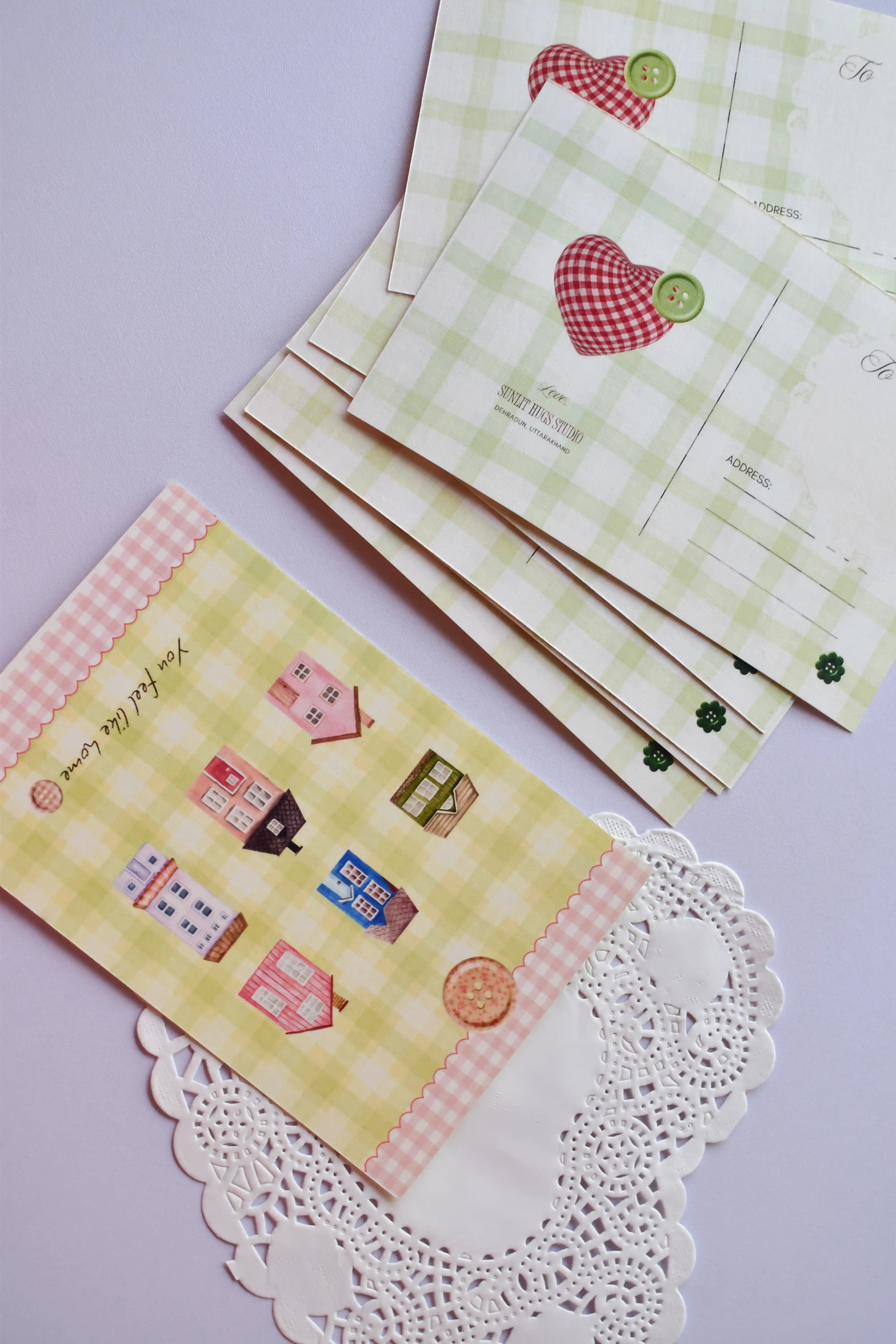 A collection of postcards showing the cozy cottage design on the bottom card, and the reverse side (or alternate card) featuring the green gingham and red patchwork heart design stacked behind it. The heart design includes spaces for address and postage.