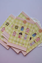 A collection of postcards showing the cozy cottage design on the bottom card, and the reverse side (or alternate card) featuring the green gingham and red patchwork heart design stacked behind it. The heart design includes spaces for address and postage.