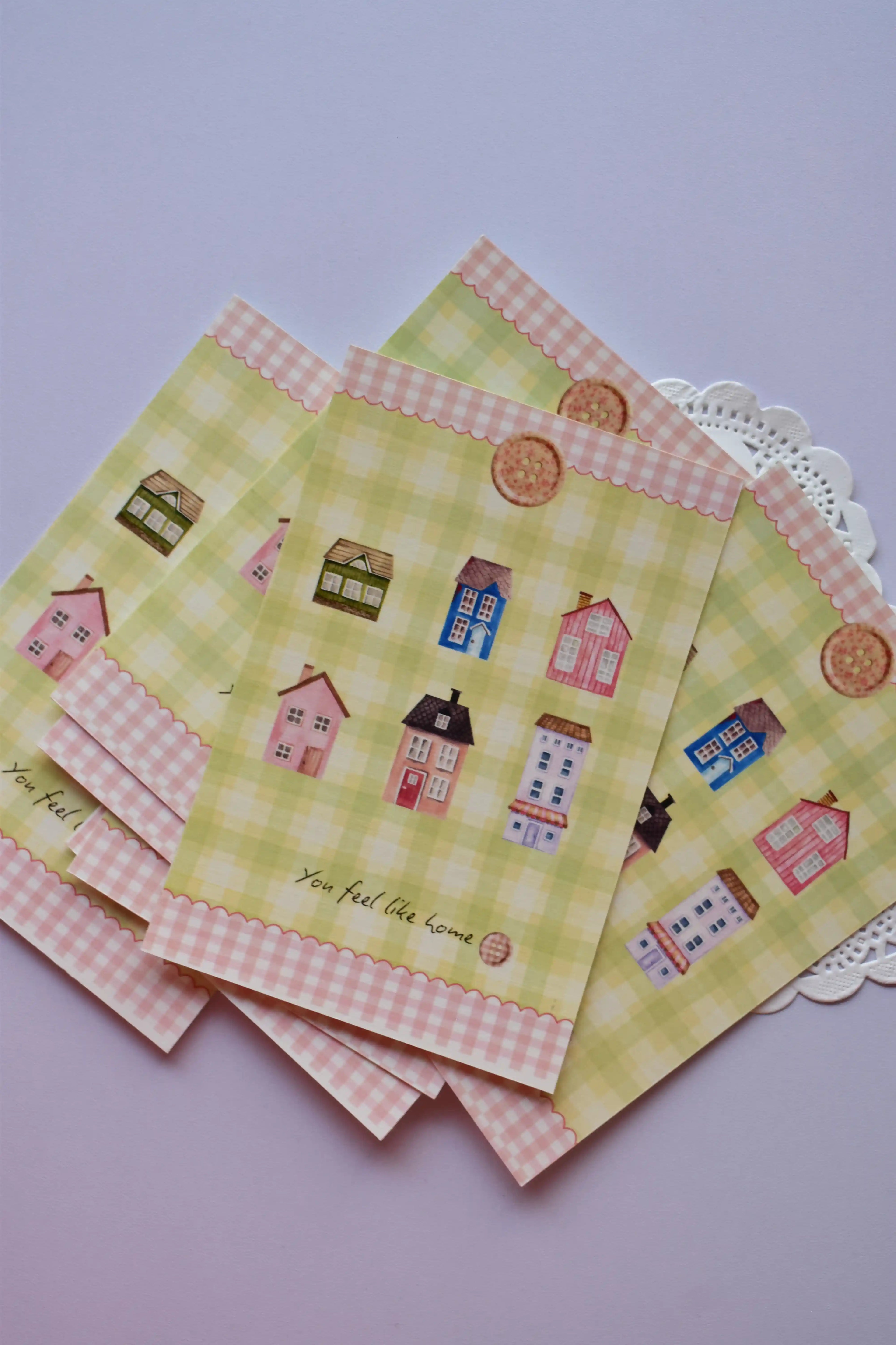A collection of postcards showing the cozy cottage design on the bottom card, and the reverse side (or alternate card) featuring the green gingham and red patchwork heart design stacked behind it. The heart design includes spaces for address and postage.