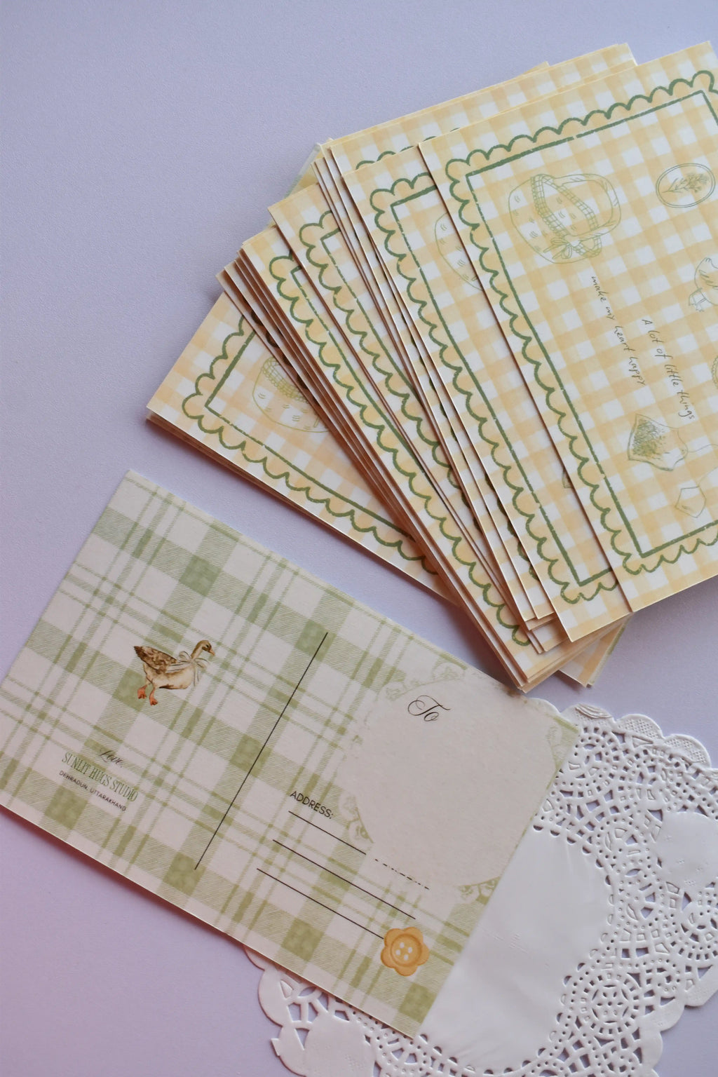 A horizontal postcard showing the green plaid reverse with the duck illustration and address lines, contrasting with a fanned stack of the yellow gingham front design featuring the 'Little Things Happy' quote.