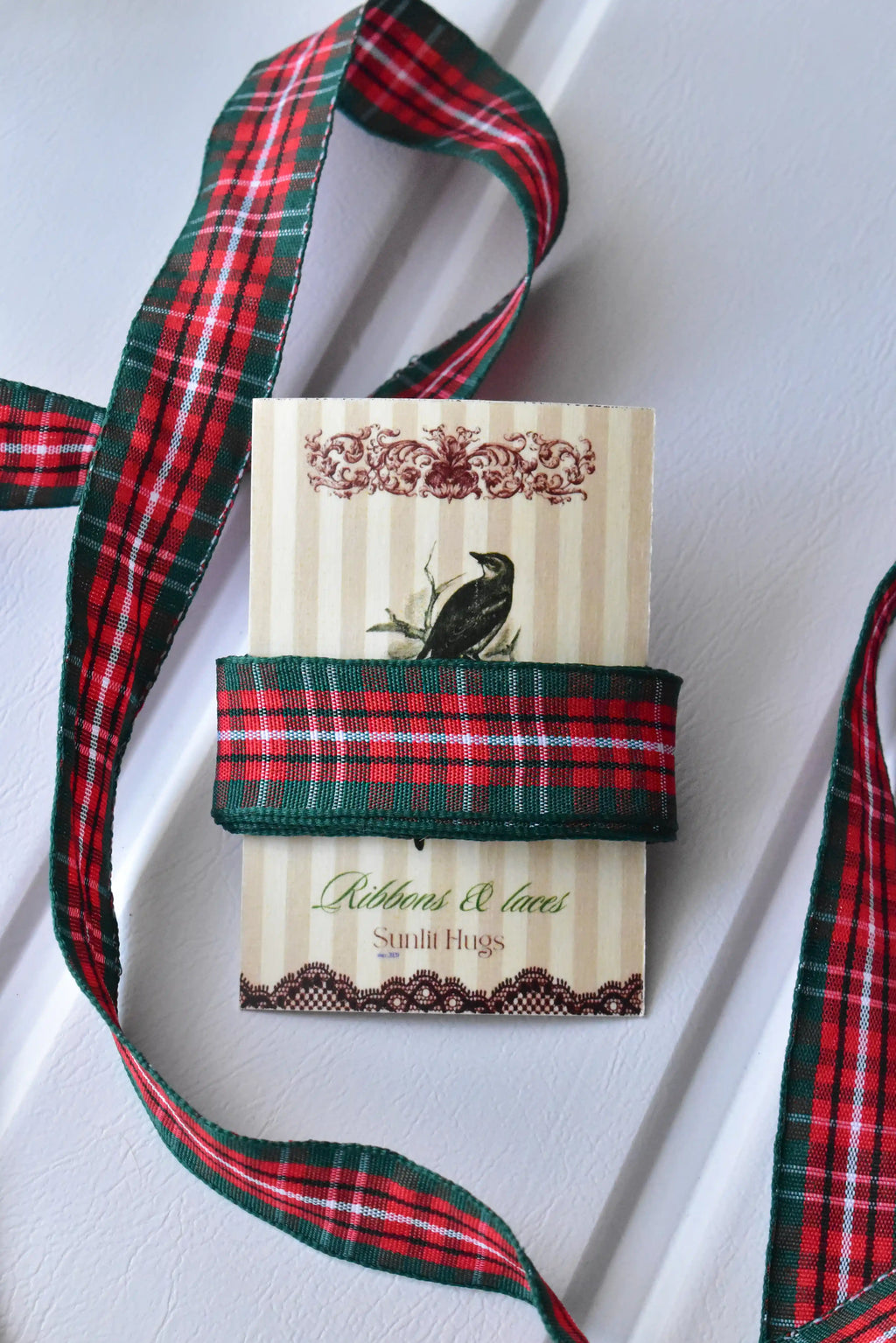This image displays the Cranberry Trail ribbon with a long, flowing strand, highlighting its red and green plaid pattern and texture. A section is also wrapped around a "Sunlit Hugs" brand tag.