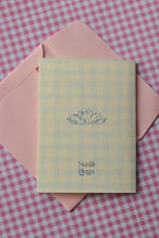 This image displays the back of the card, featuring a charming blue illustration of two swans that symbolise enduring connection, along with the Sunlit Hugs logo, sitting on a pink envelope.