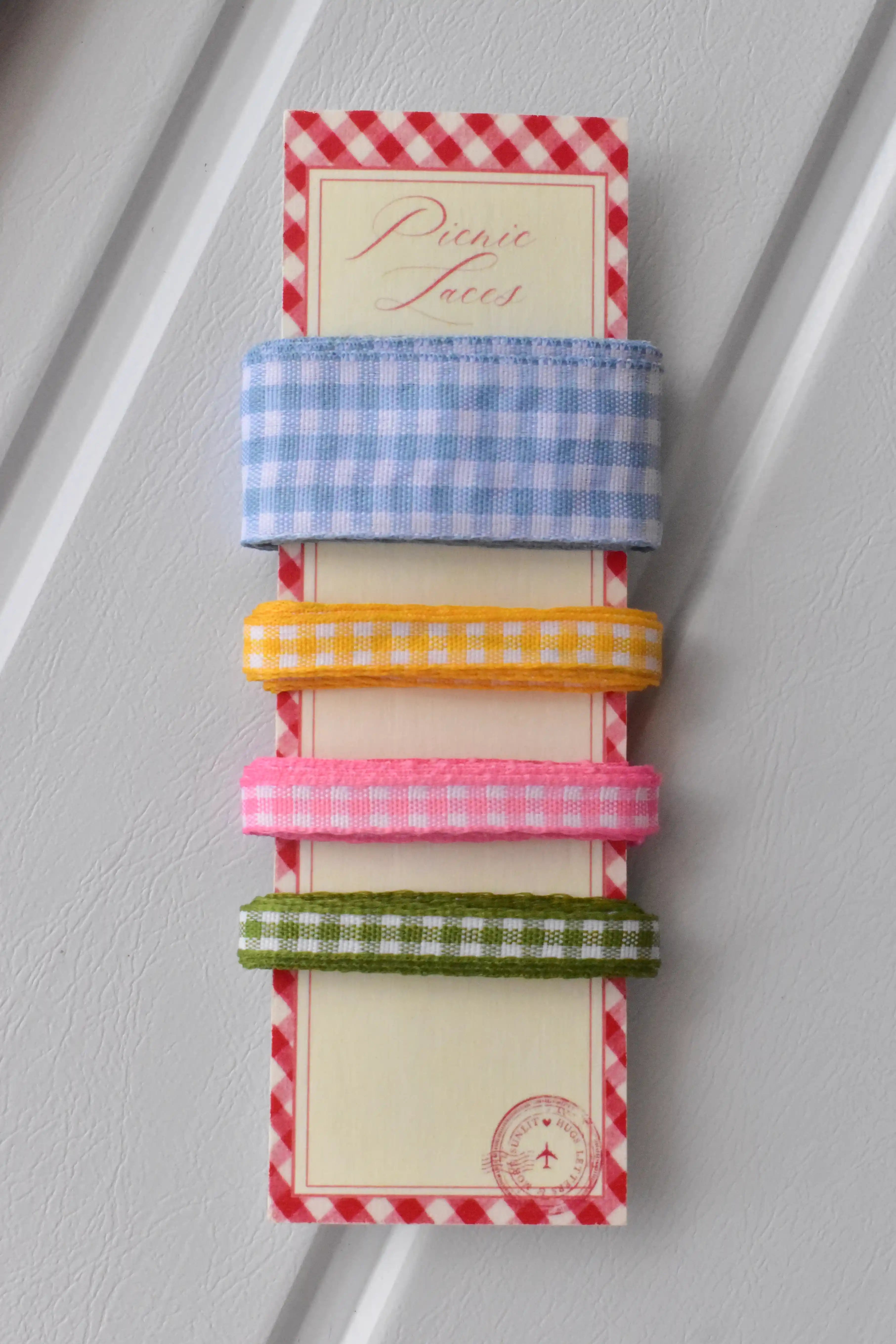 Vertical display of four narrow gingham ribbons in blue, yellow, pink, and green, set on the 'Picnic Laces' card.