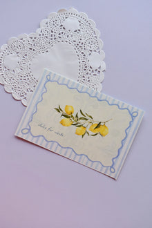 Close-up of a horizontal postcard featuring a watercolor illustration of yellow lemons on a branch. The image is framed by a decorative blue striped border and includes the text "dolce far niente." The postcard rests on a white heart-shaped paper doily.