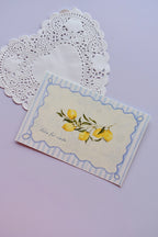 A close-up of the front of the postcard featuring bright, watercolor lemons on a branch, framed by a striped blue and scalloped border, with the phrase "dolce far niente" inscribed below.