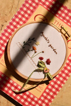 Custom hand embroidered 6-inch hoop showing the flower garden design, resting on a red and white checkered book in sunlight.