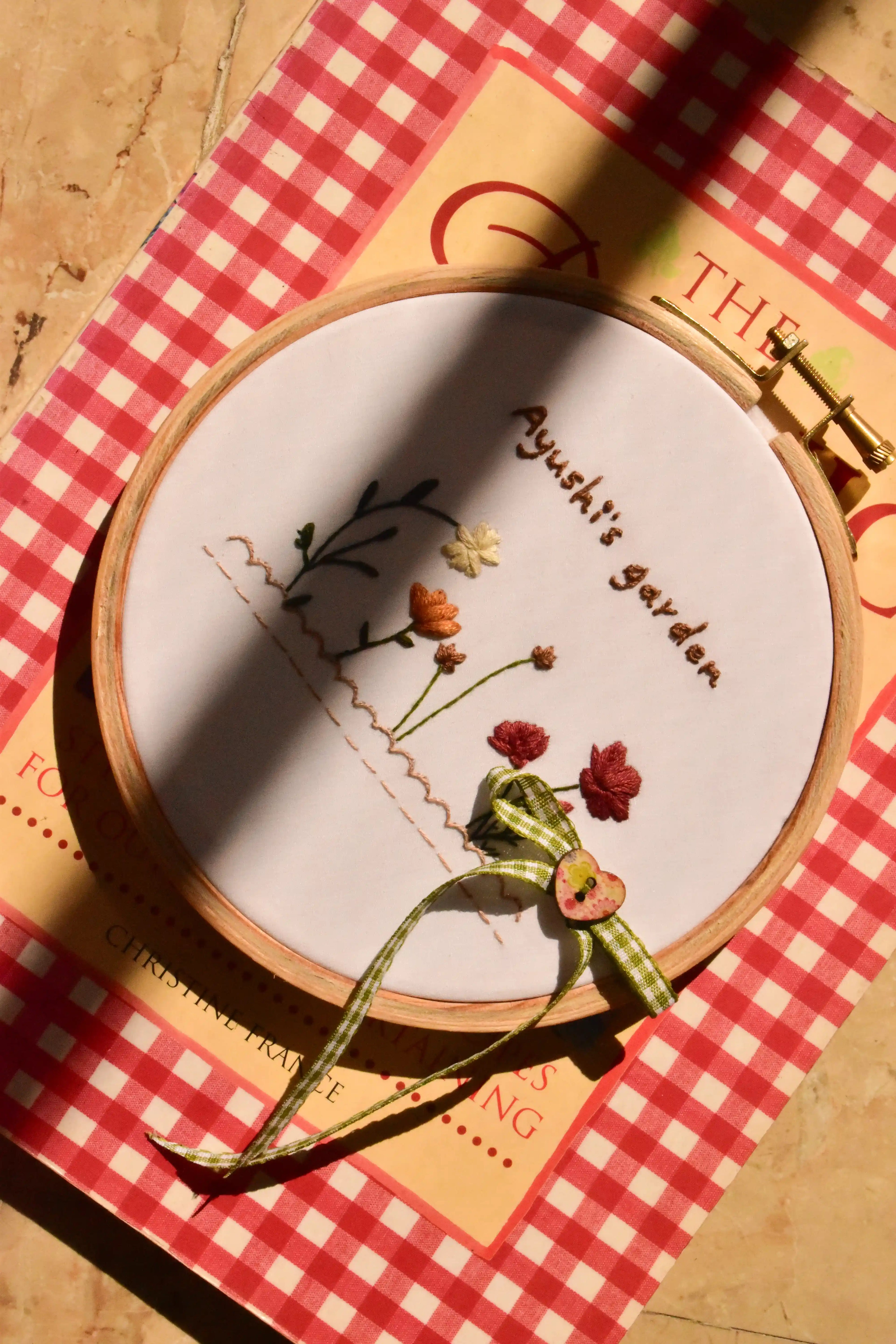 Custom hand embroidered 6-inch hoop showing the flower garden design, resting on a red and white checkered book in sunlight.
