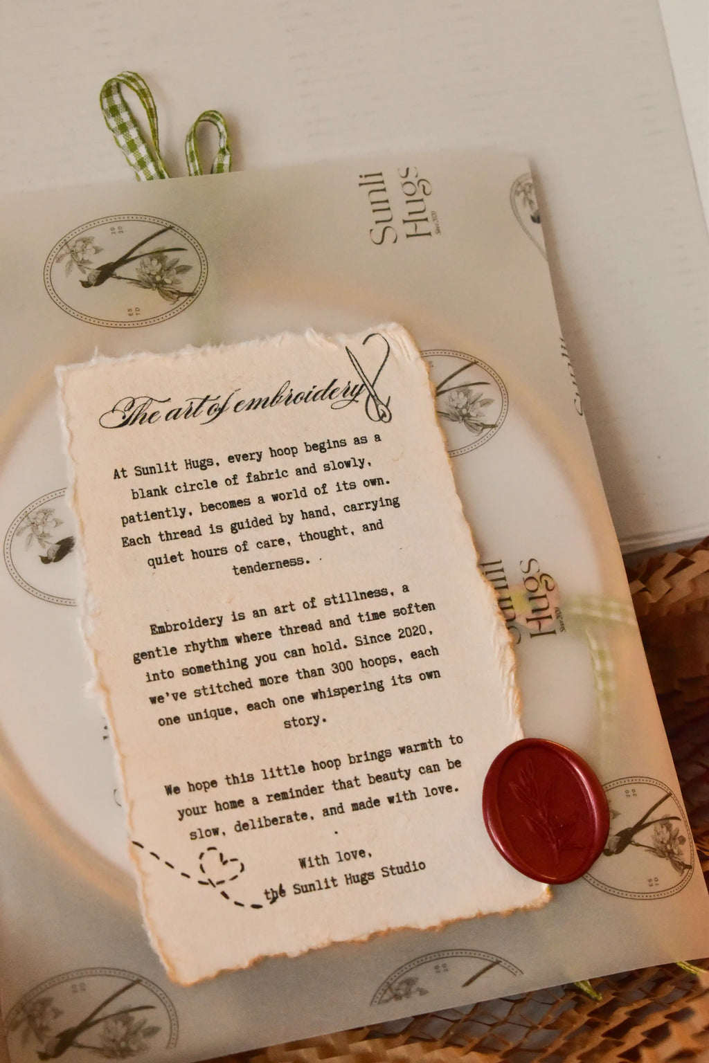 Hand embroidered hoop wrapped in Sunlit Hugs branded vellum paper, sealed with a red wax seal, and tied with a green gingham ribbon.