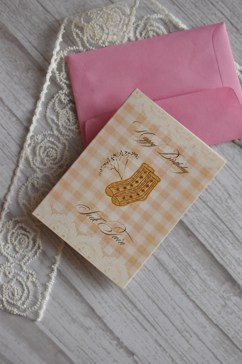 Flat lay of the Sunlit Hugs Happy Birthday Sock Twin Greeting Card resting on top of a pink envelope beside decorative white rose lace.