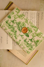 This image presents the full presentation, showing the green toile vellum envelope and golden wax seal resting on a charming stack of vintage books and papers.